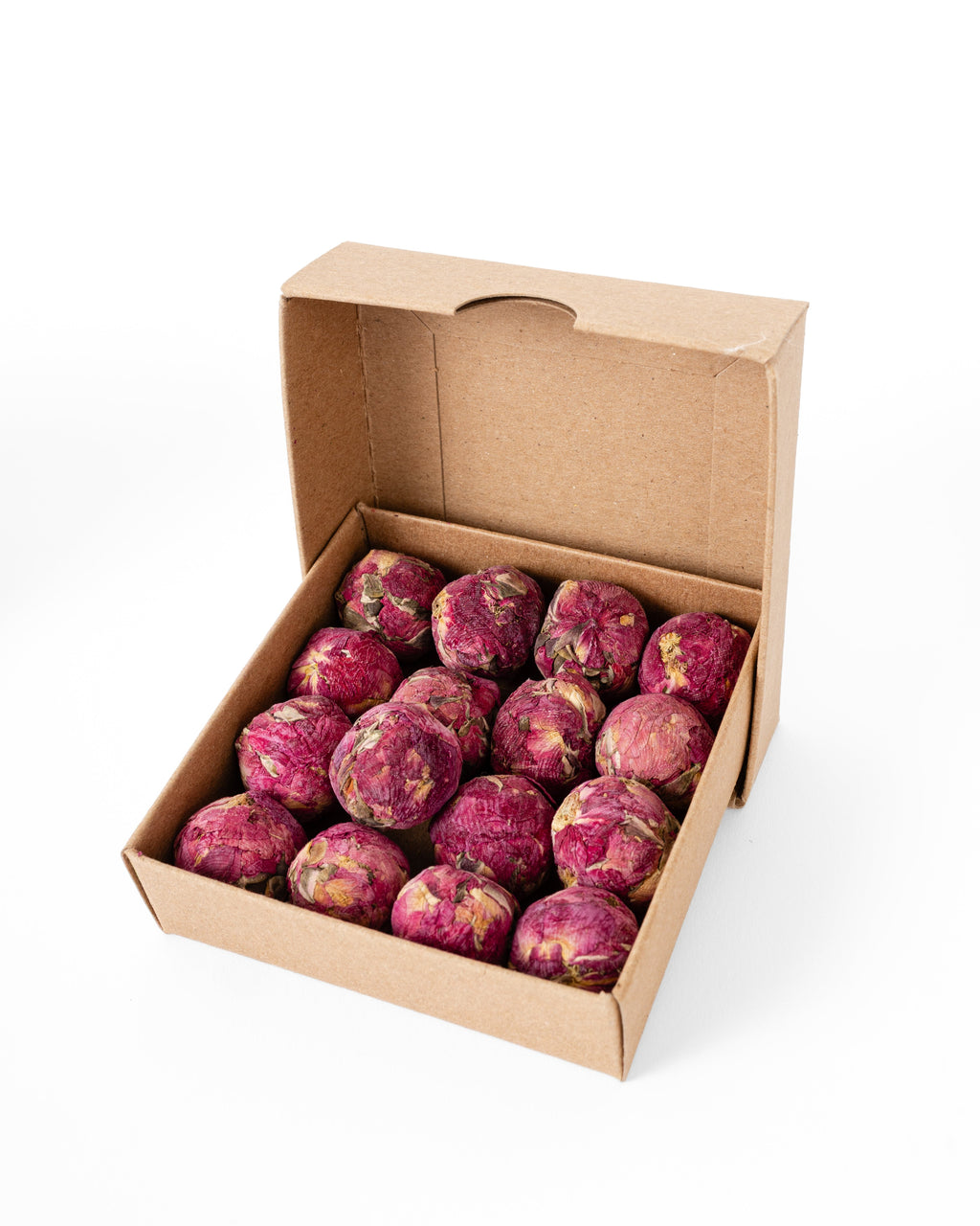 An open brown cardboard box filled with multiple small, round, dried pinkish-red rose flower balls, photographed on a clean white background.