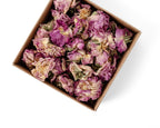 Dried pink rose buds packed in a square brown cardboard box, photographed from a top-down angle on a white background.