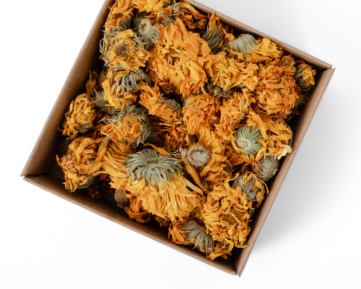 A top-down view of dried orange calendula flowers packed inside a brown cardboard box on a white background.