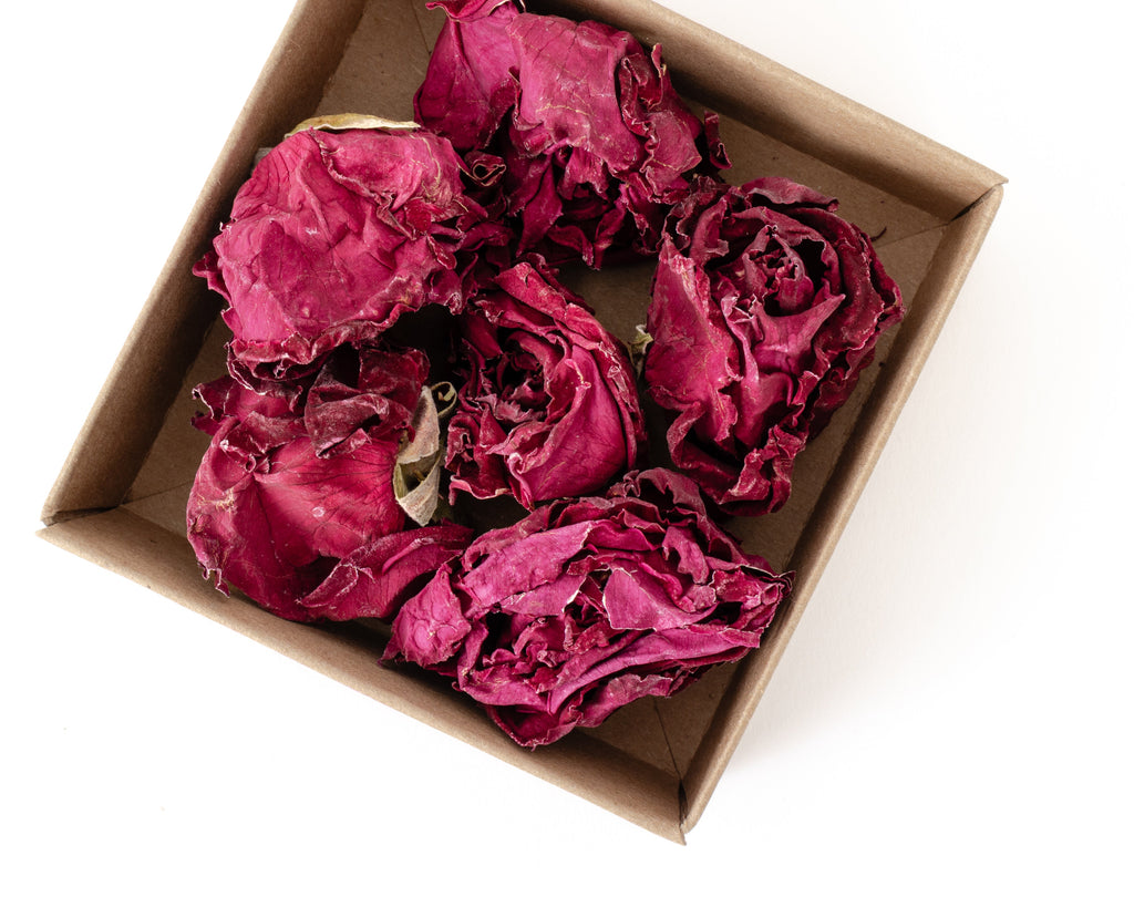 Several dried magenta-pink roses arranged inside a small brown cardboard box on a plain white background.
