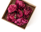 Several dried magenta-pink roses arranged inside a small brown cardboard box on a plain white background.