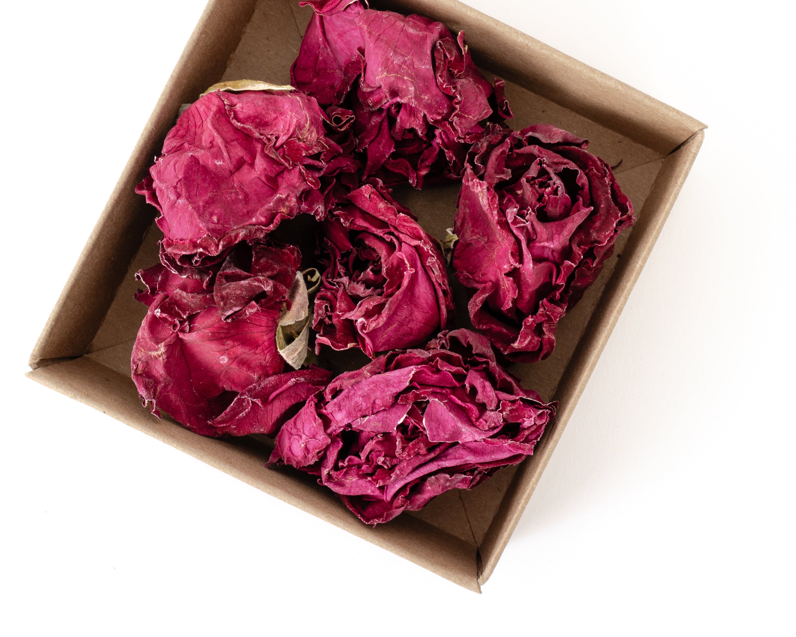 Several dried magenta-pink roses arranged inside a small brown cardboard box on a plain white background.