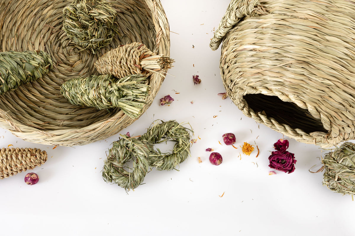 Natural handmade bunny bed and hidey house on a white background, with dried flowers and chew toys arranged around them.