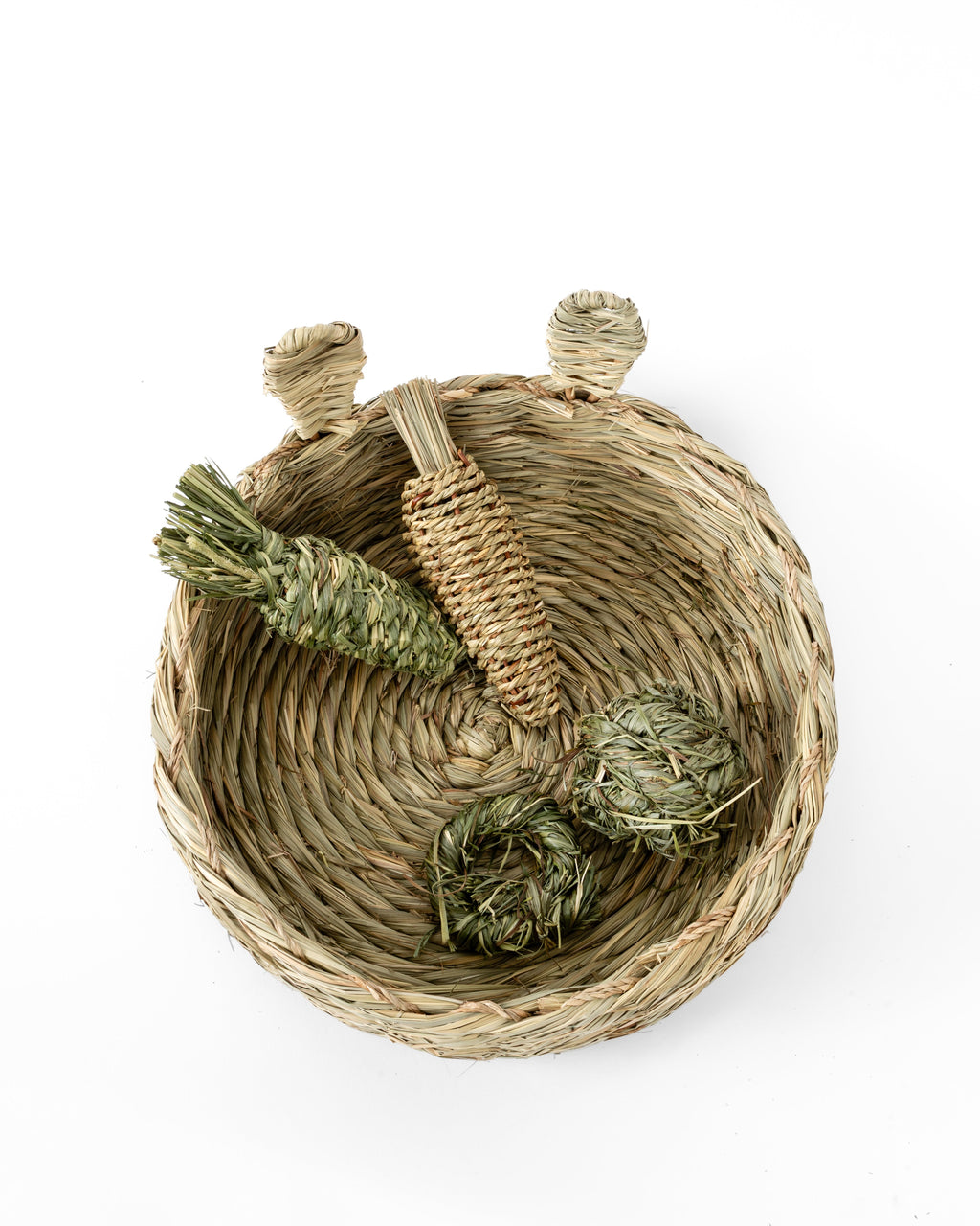 A natural woven thatch rabbit bed with, shown from above on a white background. Inside the basket are several small handwoven toys made from timothy grass or reed.