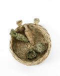 A natural woven thatch rabbit bed with, shown from above on a white background. Inside the basket are several small handwoven toys made from timothy grass or reed.